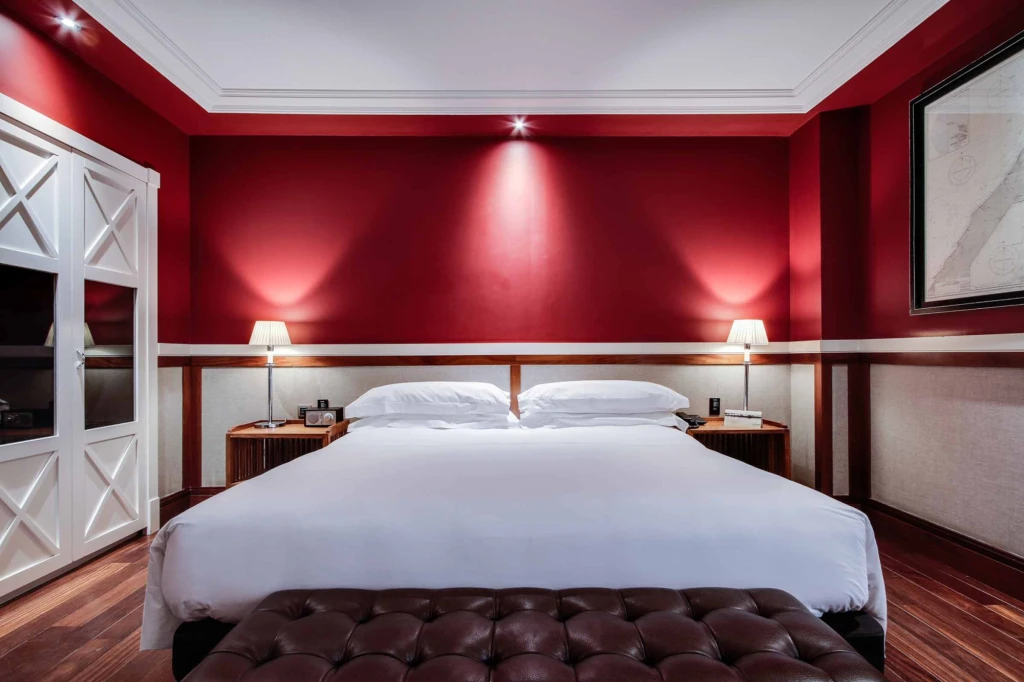 A double bed with an upholstered headboard, two bedside tables, and lamps is shown in a room with red walls.