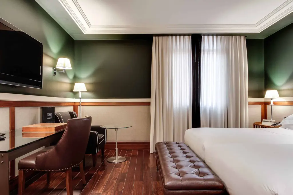La imagen muestra una habitación con cama, escritorio, sillón y cortinas junto a la ventana.