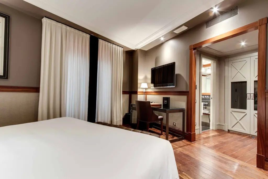 Das Zimmer verfügt über ein Bett, Vorhänge, einen Fernseher und einen Schreibtisch neben einer Innentür.