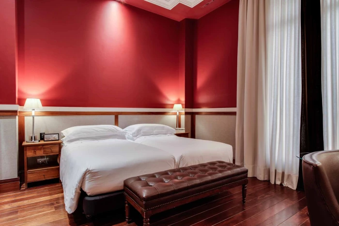 The image shows a double bed with white linens, bedside tables, and curtains next to a red wall.