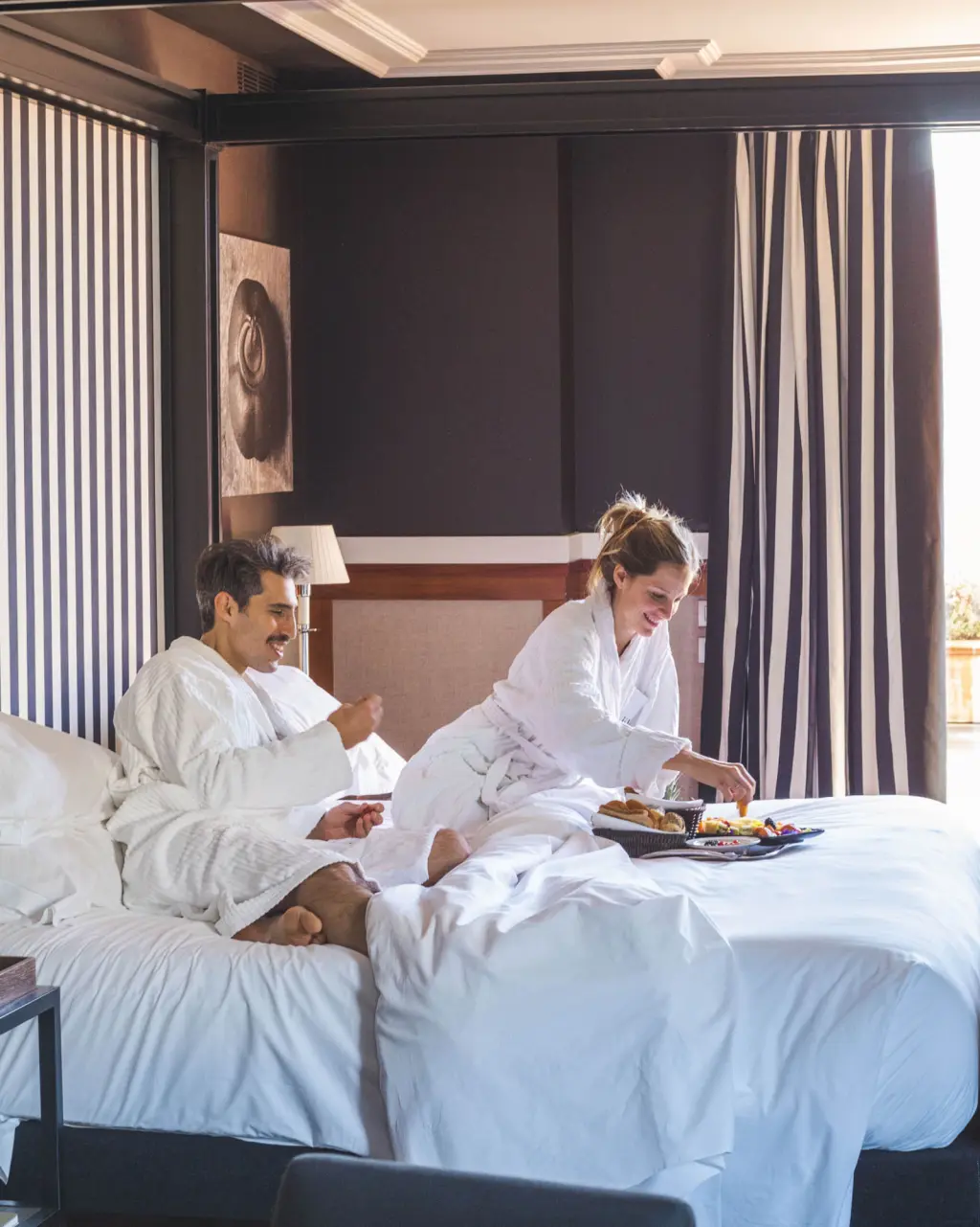 Deux personnes sont assises sur un lit avec des draps blancs et des rideaux tout autour.
