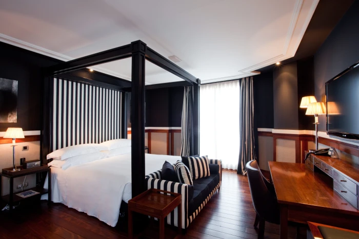 The image shows a junior suite with a bed, striped armchair, desk, and floor lamps.