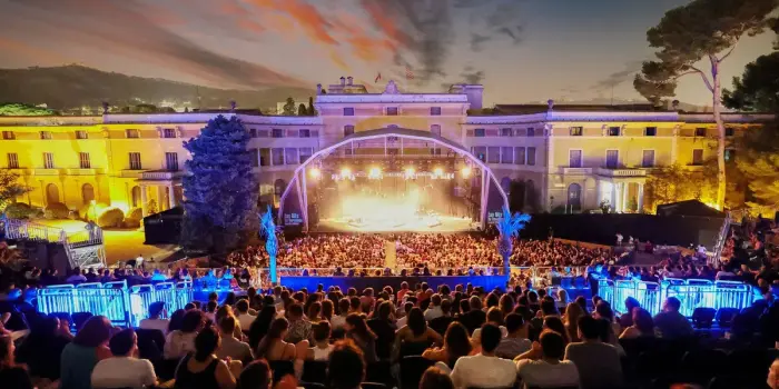 La imagen muestra un escenario iluminado con un público numeroso frente a un edificio iluminado.