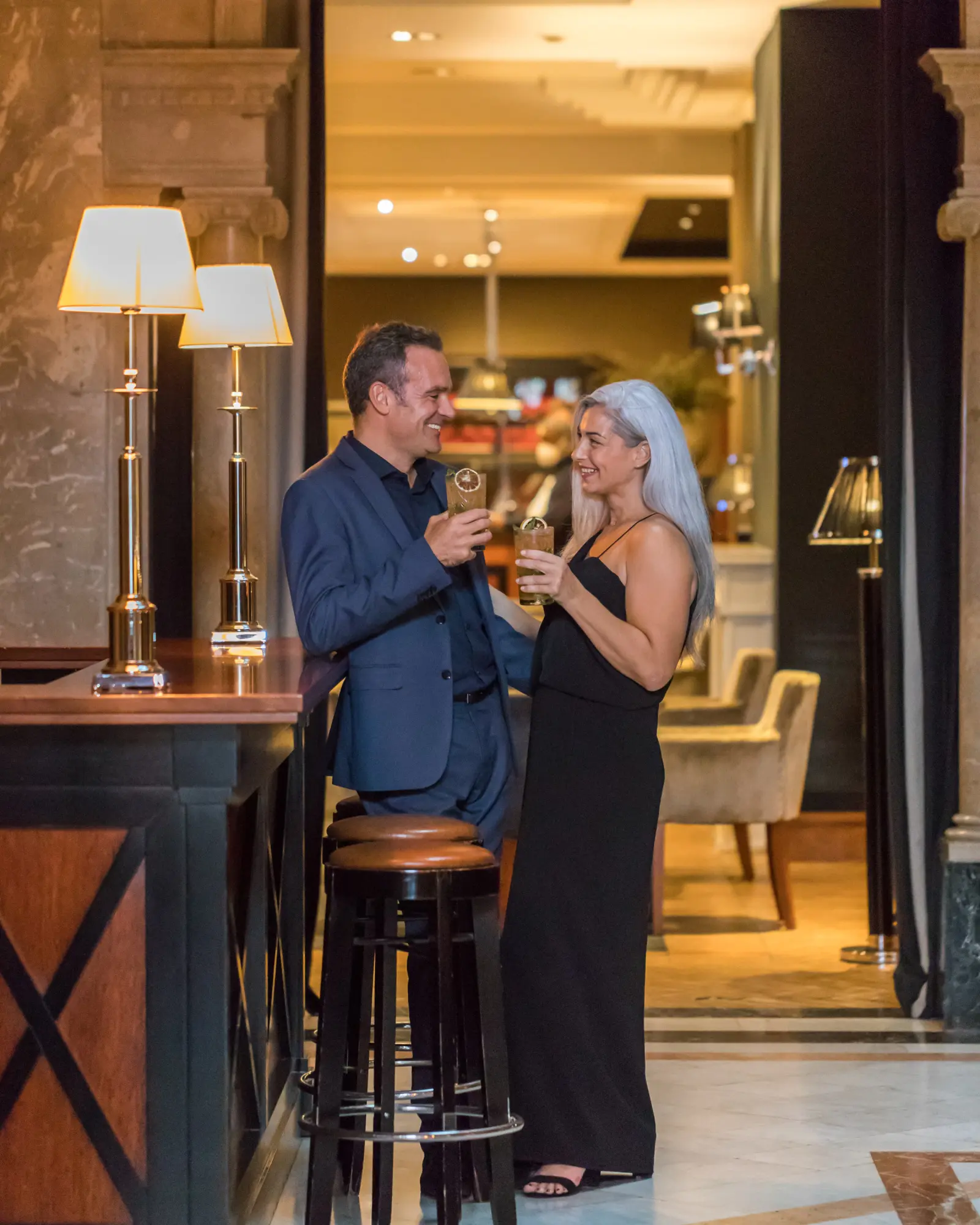 A couple toasts with glasses in a hotel lobby with tables and lit lamps.