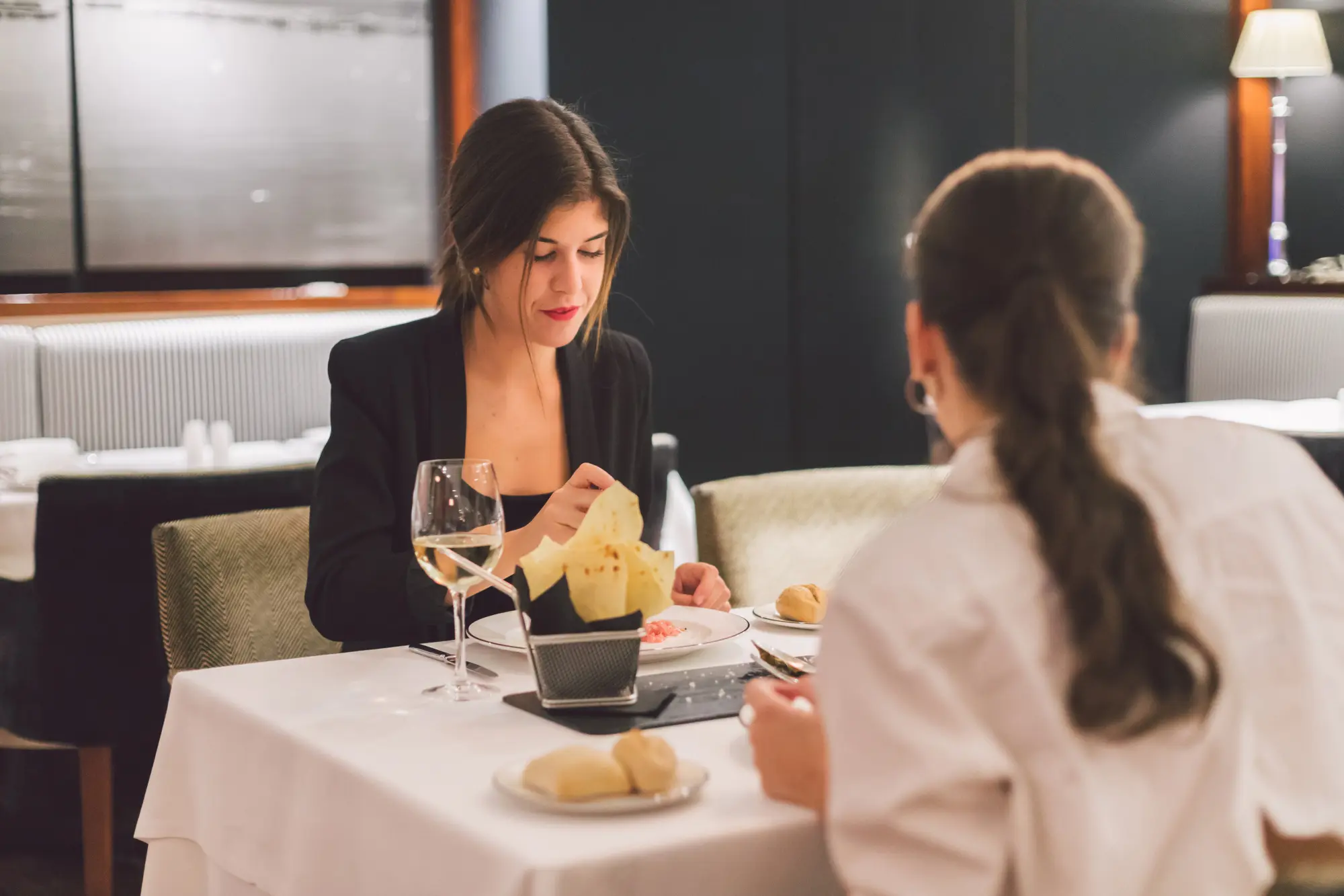 A woman and another person dine at a table with glasses and plates on a white tablecloth.