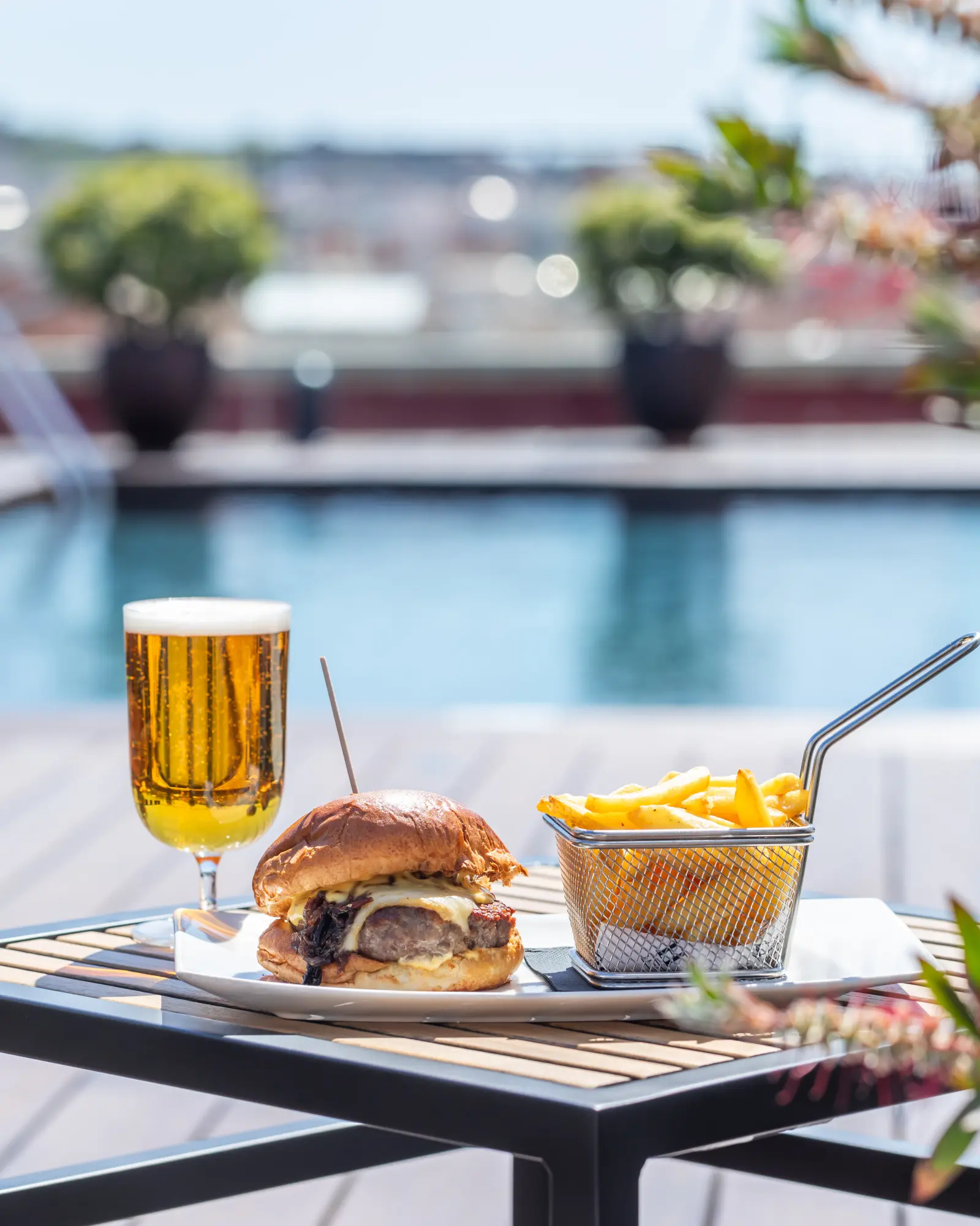 Une table en terrasse extérieure est dressée avec un burger, des frites, une carafe de bière et des verres.