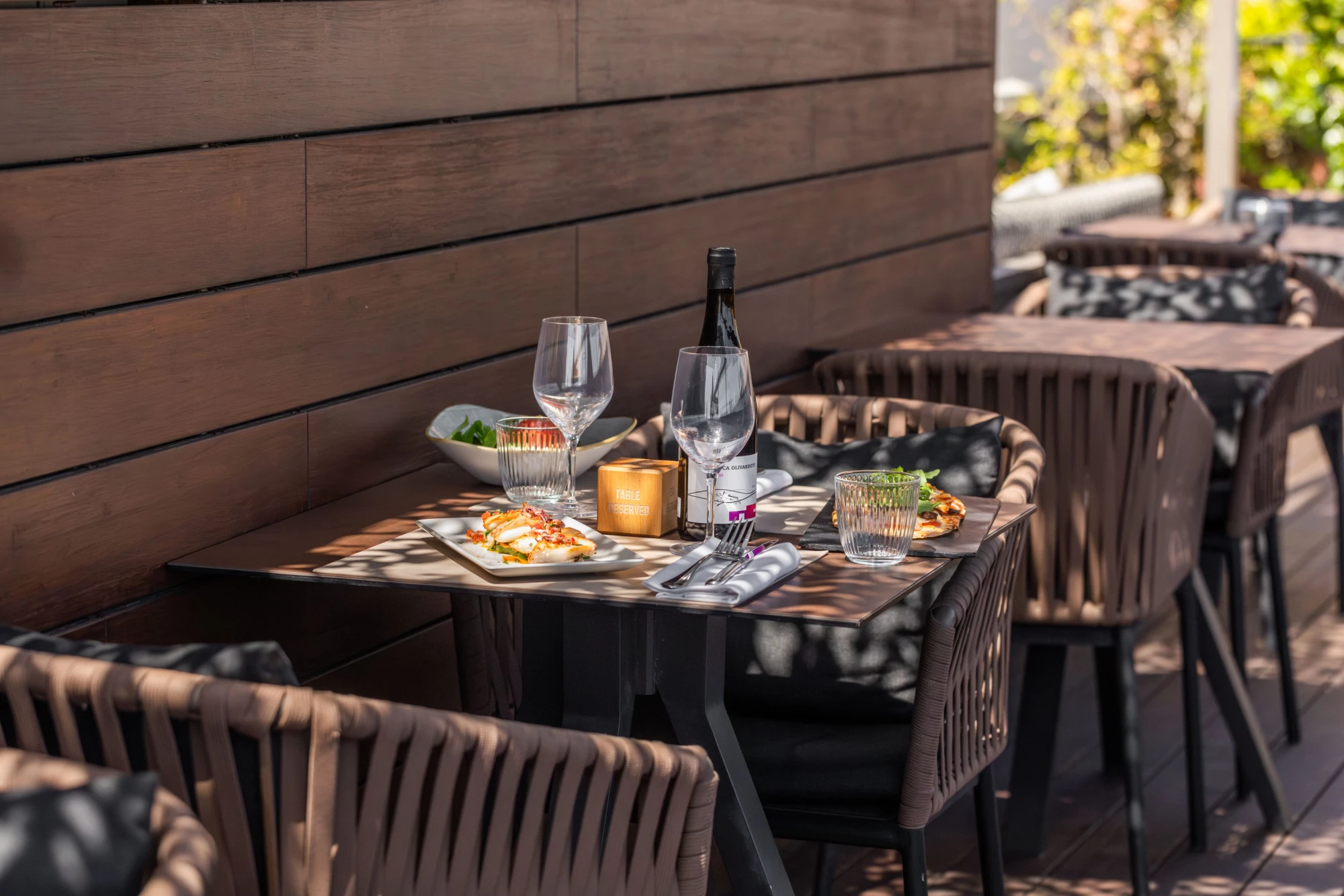 Se ve una mesa con copas y platos sobre un mantel, con sillas alrededor y un muro de fondo.