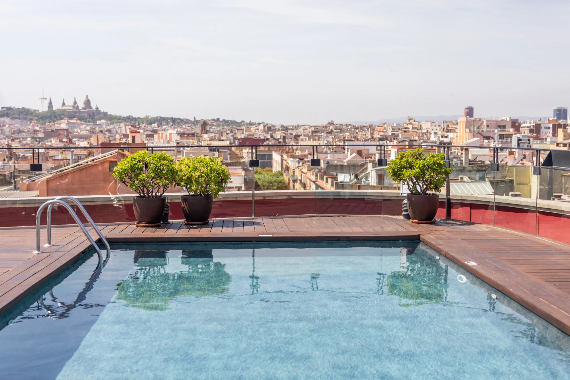 Se ve una piscina en una terraza con barandillas y macetas, con la ciudad al fondo.