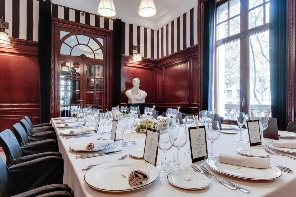 La imagen muestra un salón con paredes a rayas, una mesa preparada con copas y tarjetas de menú, y ventanas altas.