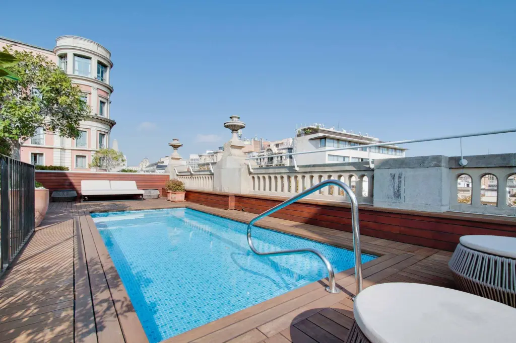 Se ve una piscina en una terraza con barandilla de metal y tumbona junto a un edificio de varios pisos.