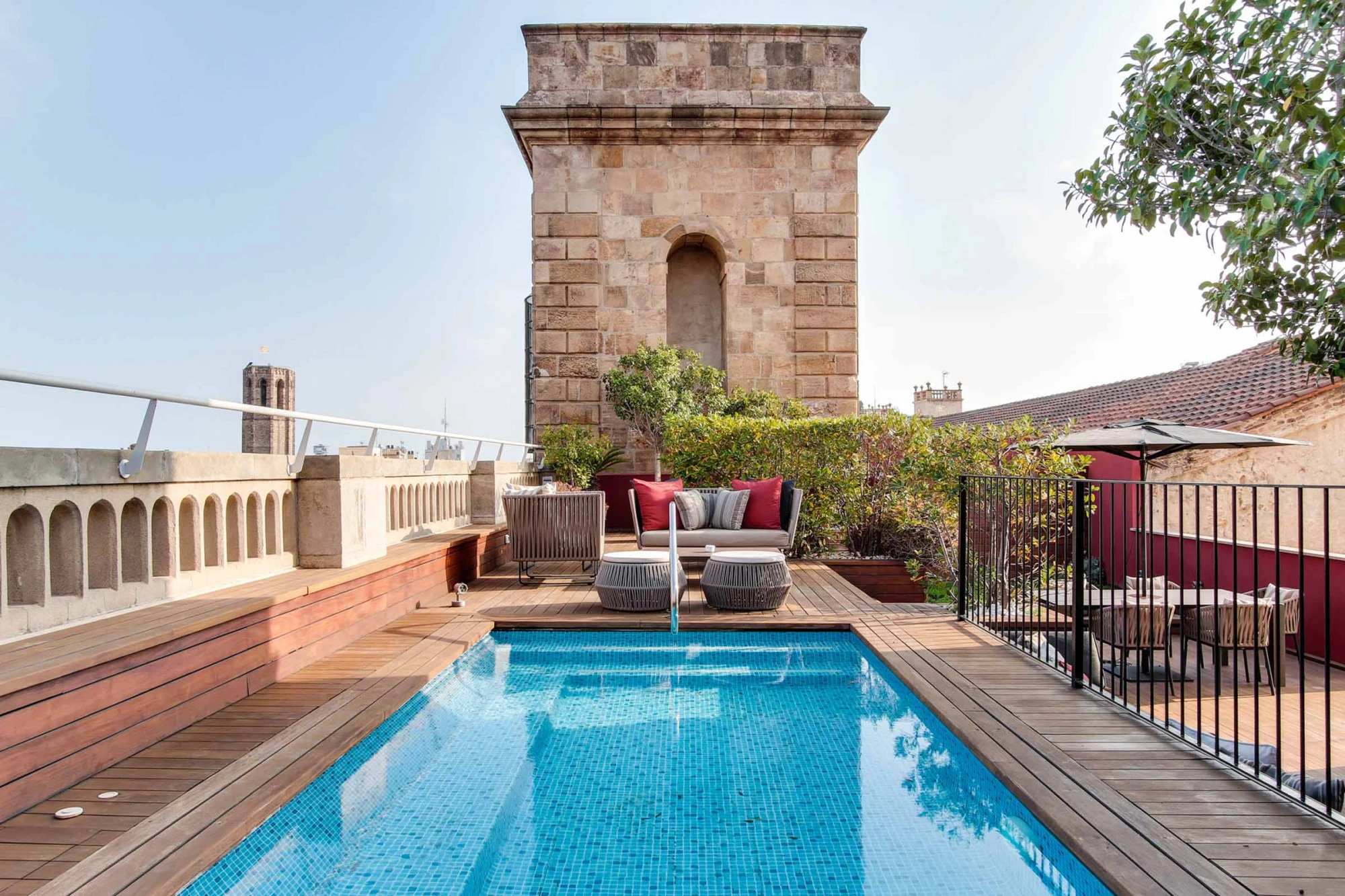 Se ve una piscina azul en una terraza con tumbonas, barandillas y plantas alrededor.