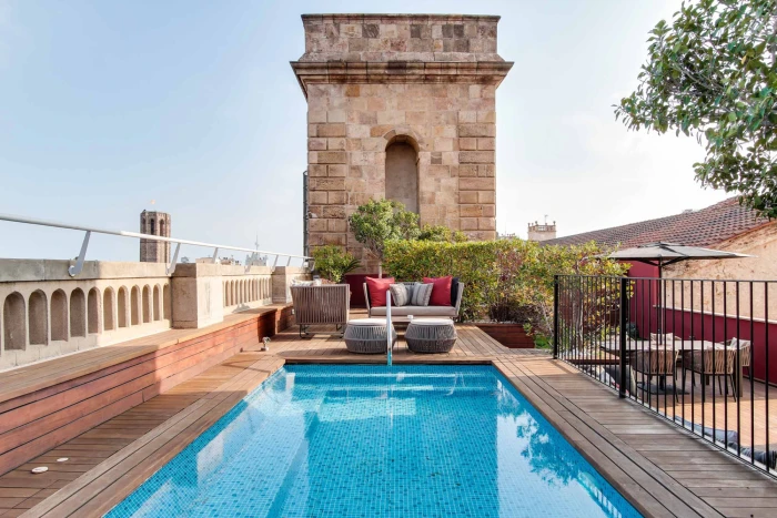 A blue pool is visible on a terrace with loungers, railings, and plants around it.
