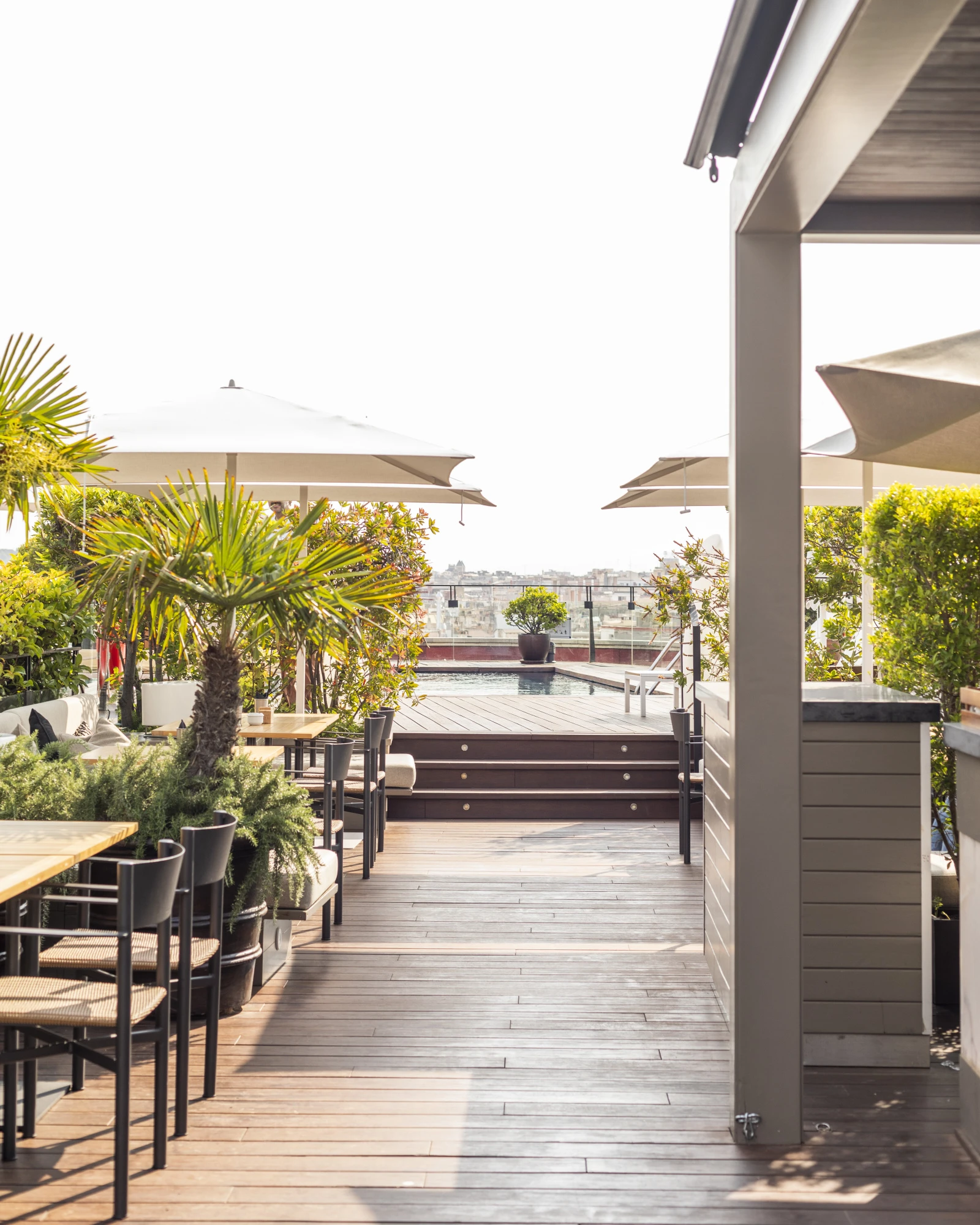 Man sieht eine Terrasse mit Tischen und Stühlen, Topfpflanzen und einem Geländer mit Blick auf die Stadt.