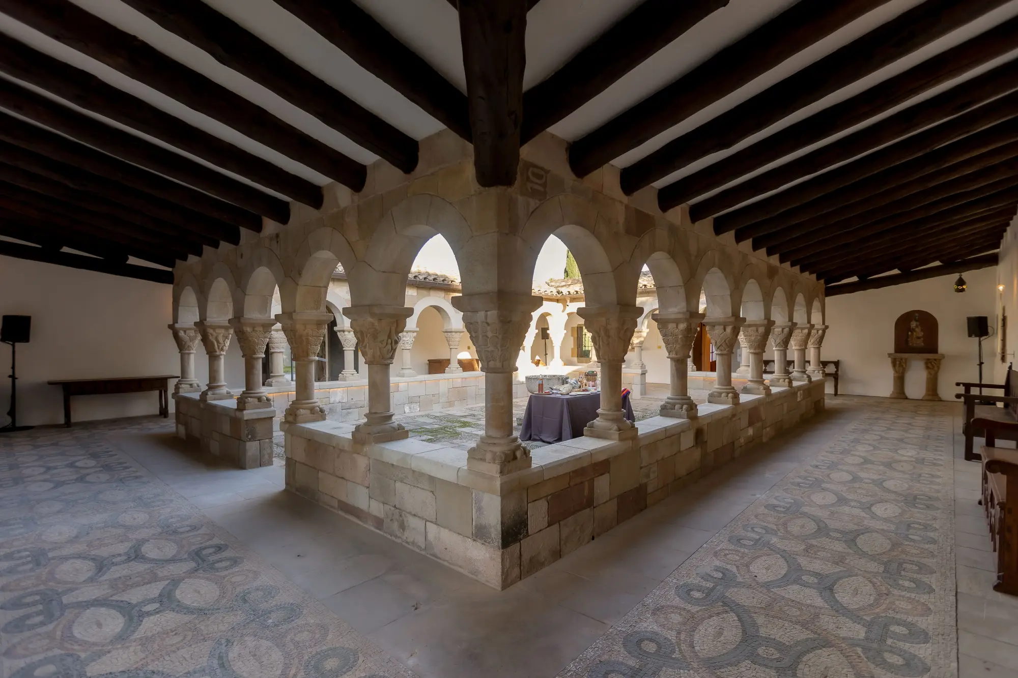 Se ve una galería interior con arcos de piedra, columnas y un techo de vigas de madera.