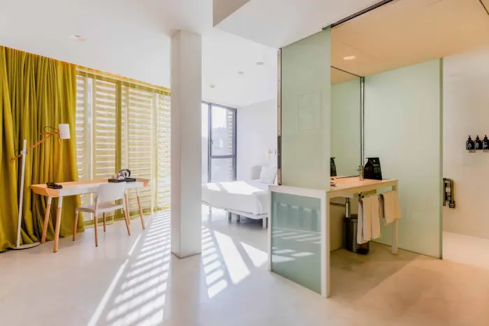 Se ve una sala luminosa con sofá, escritorio y una mesa junto a una ventana con cortinas.