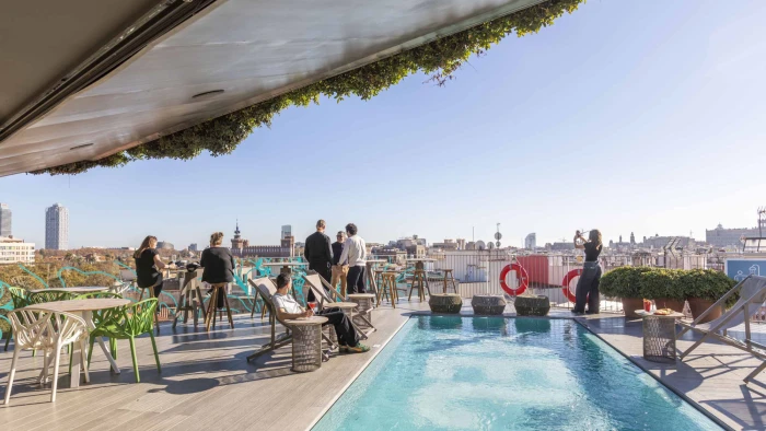 Varias personas descansan junto a una piscina en una terraza con vistas a un paisaje urbano.