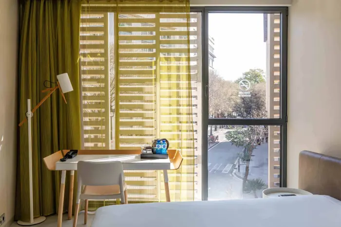 Se ve una cama, un escritorio con silla y una ventana con persianas en la habitación.