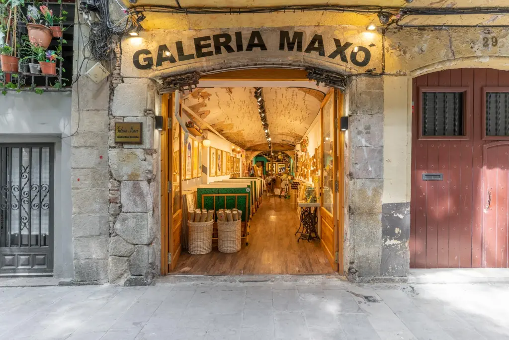 Se ve la entrada de una galería con letrero “GALErIA MAXO” y un pasillo interior con mesas y sillas.