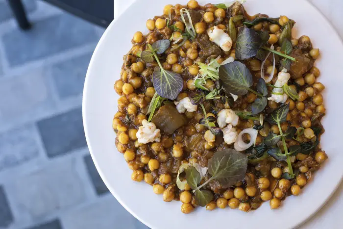 Sirve un plato blanco con garbanzos y hojas verdes sobre una mesa de piedra.