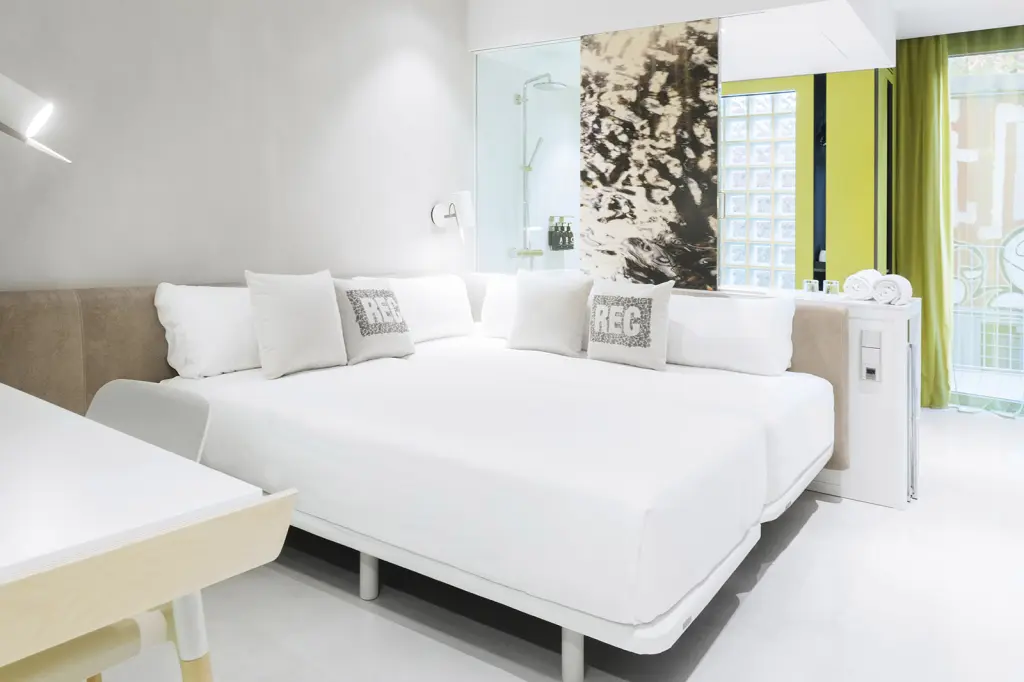 La imagen muestra una cama doble blanca con almohadas y una ventana con cortinas verdes.