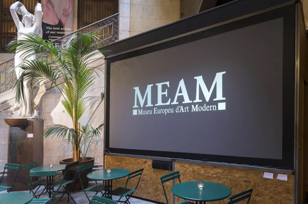 Se ve una pantalla con el texto “MEAM” y una zona exterior con mesas y sillas junto a una pared de ladrillo.