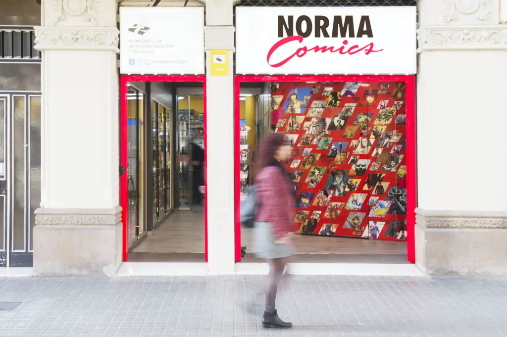 Una persona camina frente a una entrada con un escaparate rojo y el letrero “NORMA Comics”.