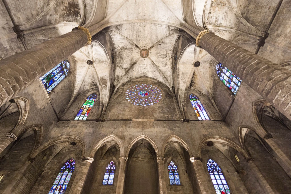 Se ve el interior de una iglesia con bóveda de piedra y vitrales en las ventanas altas.