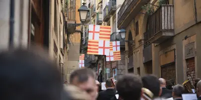 Personas observan un pasacalles con banderas colgadas en una calle urbana estrecha.
