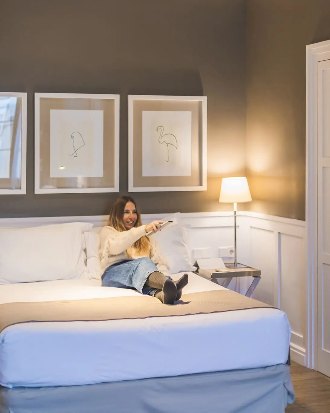 Una mujer se sienta sobre una cama con ropa de cama blanca y una lámpara de mesa junto a la pared.