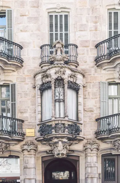 Se ve una fachada de piedra con ventanas con persianas y un balcón de hierro forjado con decoración escultórica.