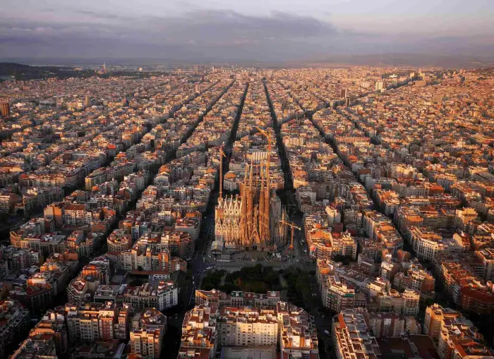 Es veu un paisatge urbà de l’Eixample amb carrers llargs i edificis densos sota un cel ennuvolat.