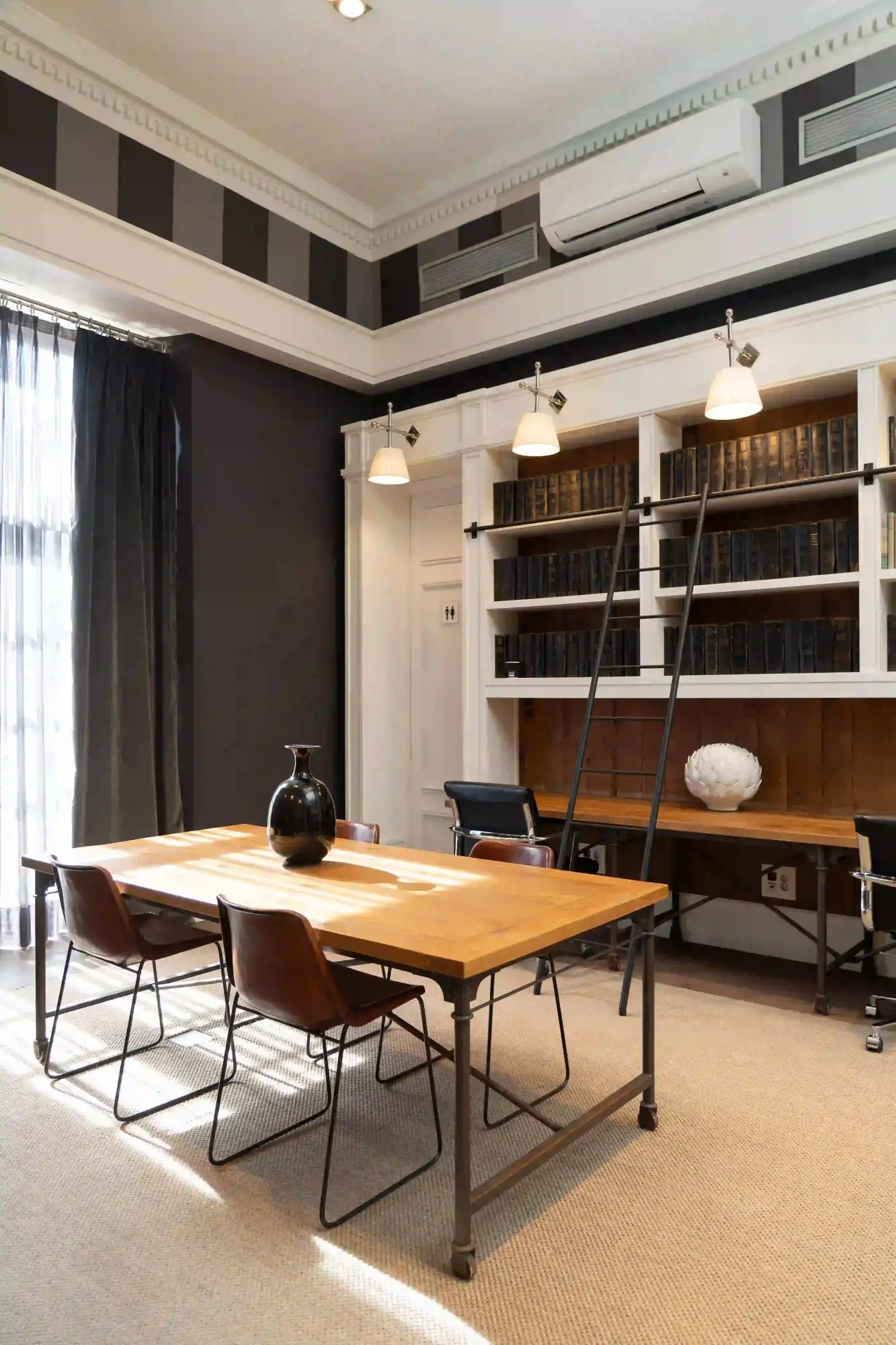 Si vede una biblioteca con tavolo in legno, sedie in metallo e librerie a parete piene di libri.