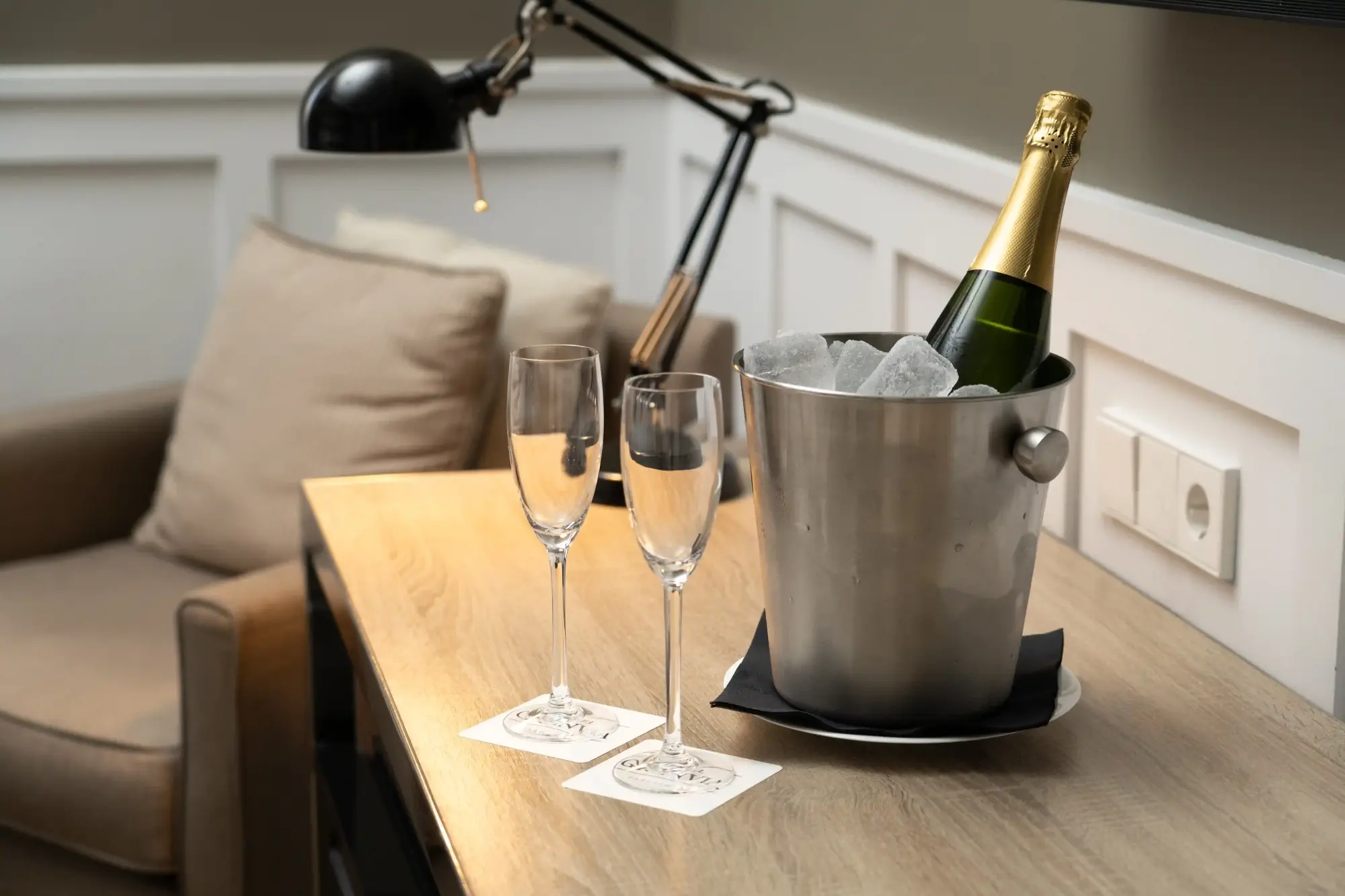 Se ve una mesa con una cubitera metálica con botellas y dos copas sobre un sofá tapizado.