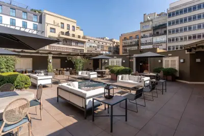Se ve una terraza amplia con mesas, sillas y sombrillas, rodeada de vegetación y edificios urbanos al fondo.