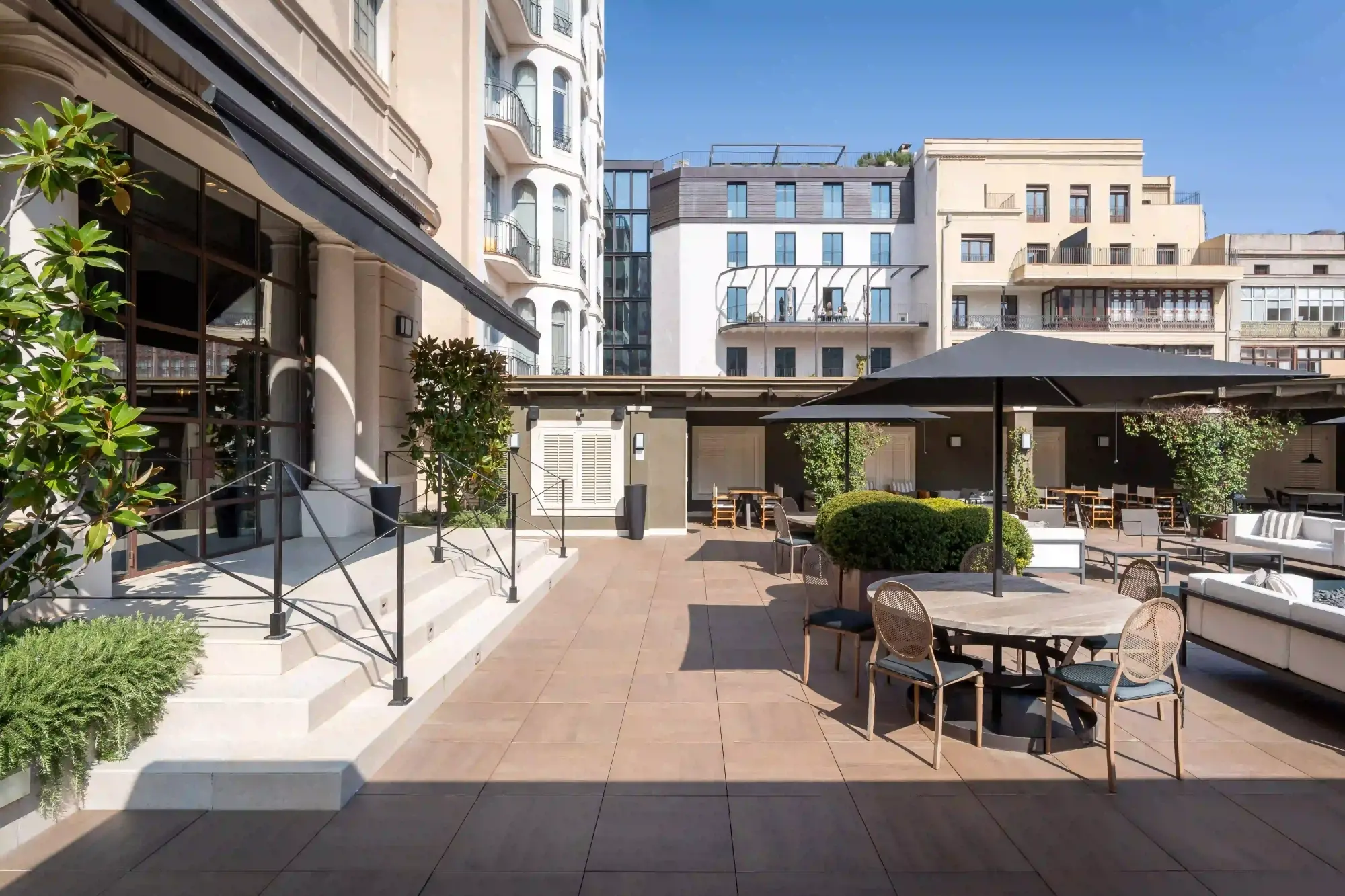 An outdoor terrace is shown with tables and chairs, planters and umbrellas beside a building.