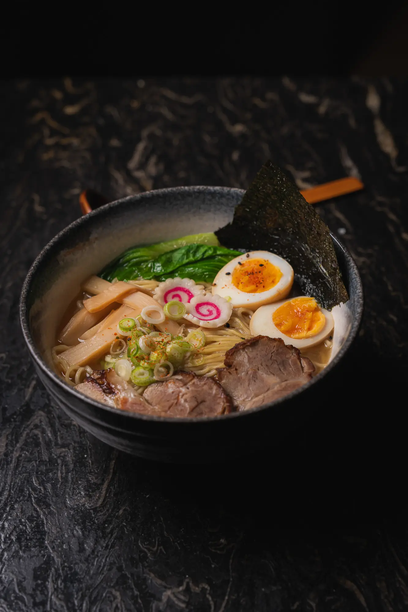 Se ve un tazón de ramen oscuro con rodajas de huevo, cebollín y fideos sobre un fondo oscuro.