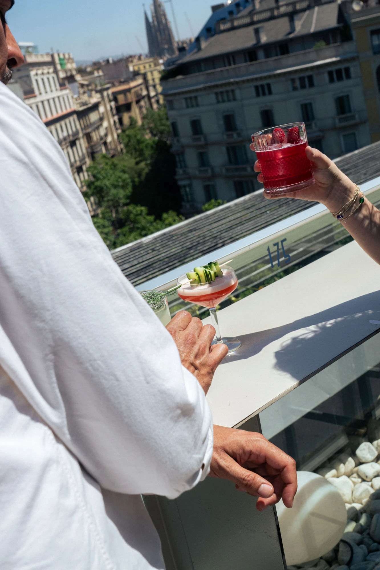 Dos personas brindan con vasos de bebida sobre una mesa en un balcón con vista urbana.