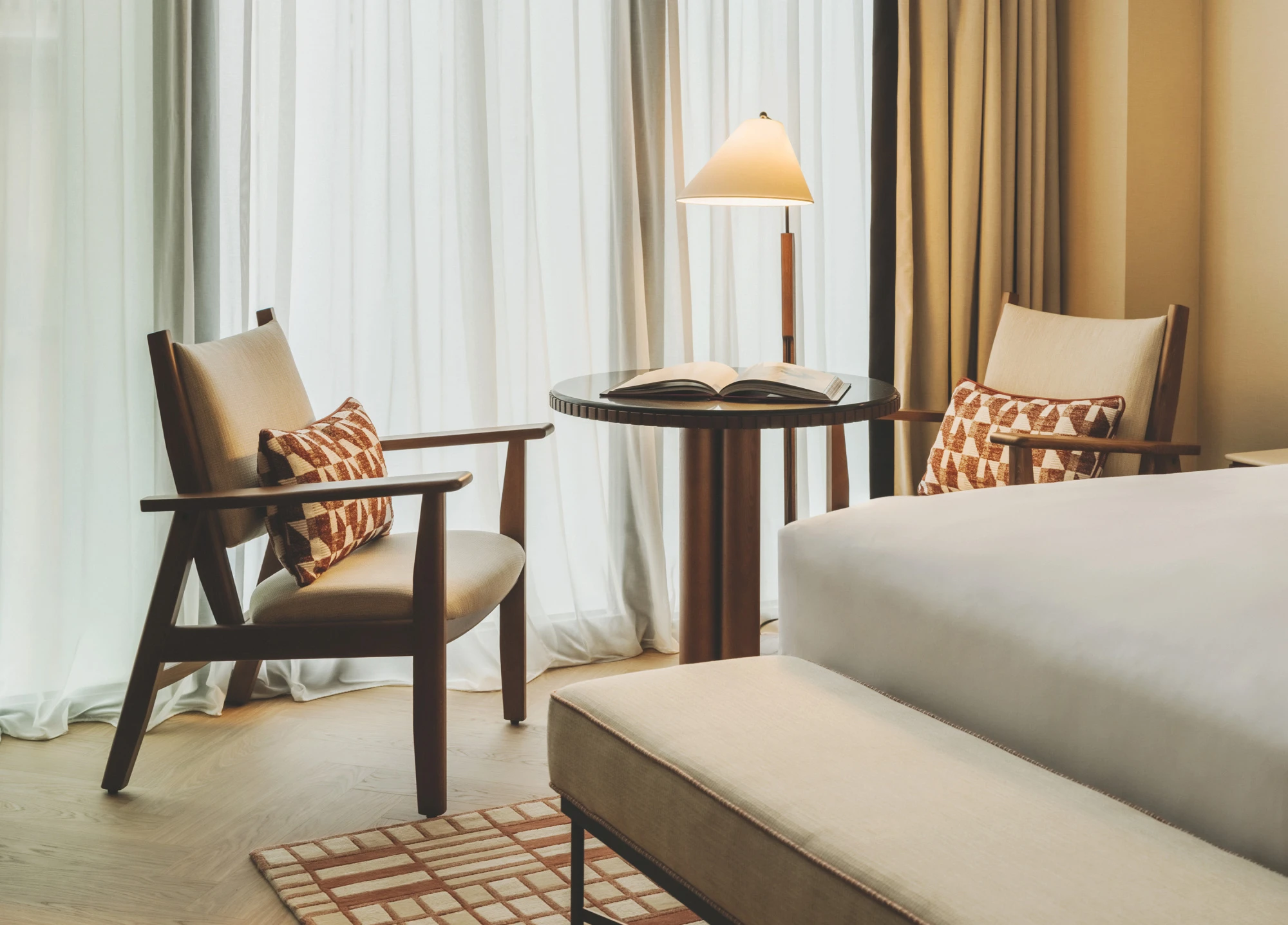 Se ve una cama, dos sillones, una mesa auxiliar redonda y una lámpara sobre una mesita junto a cortinas.