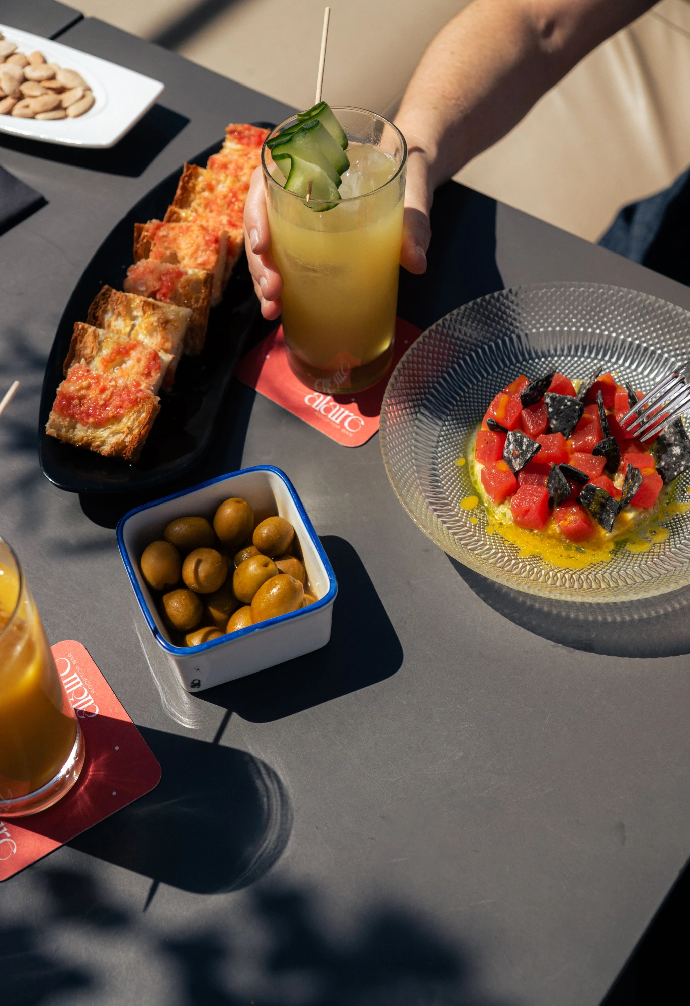 Se ve una mesa con bebidas en vasos, aceitunas en un recipiente y platos con comida.