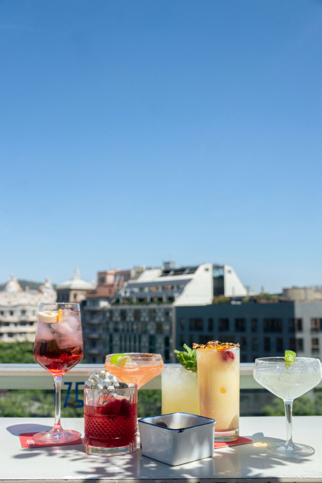 Se ven varios cócteles en vasos sobre una mesa en una terraza con edificios al fondo.