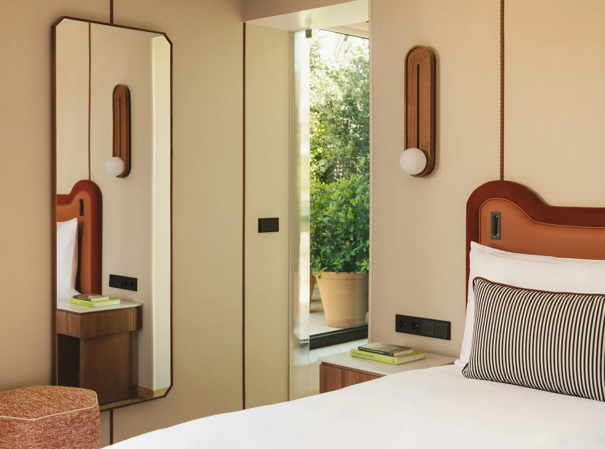 Muestra una cama con cabecero y cojines, un espejo y una consola junto a una puerta abierta hacia el exterior.
