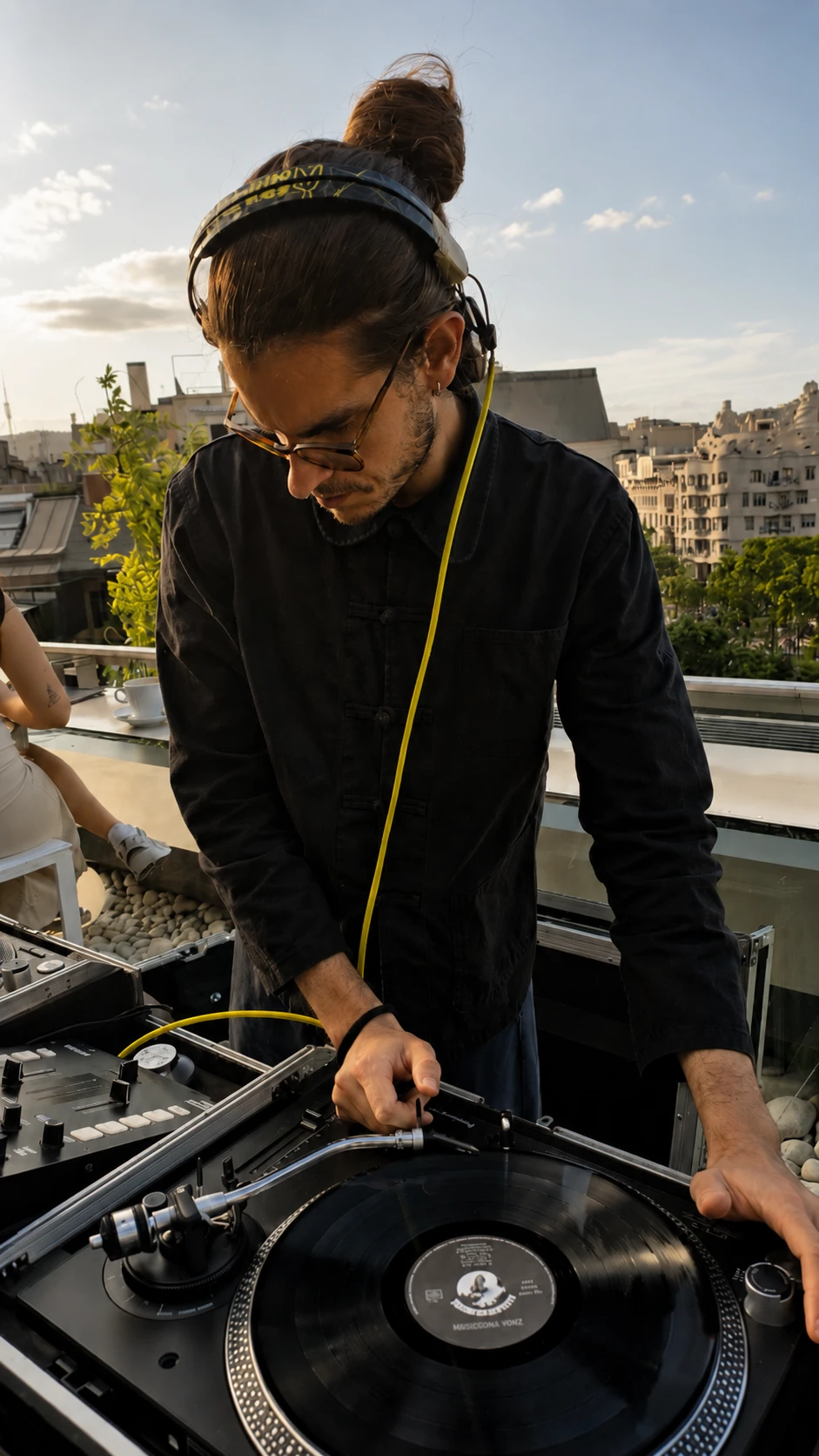 Un DJ ajusta un tocadiscos en una cabina sobre una terraza con vista urbana.