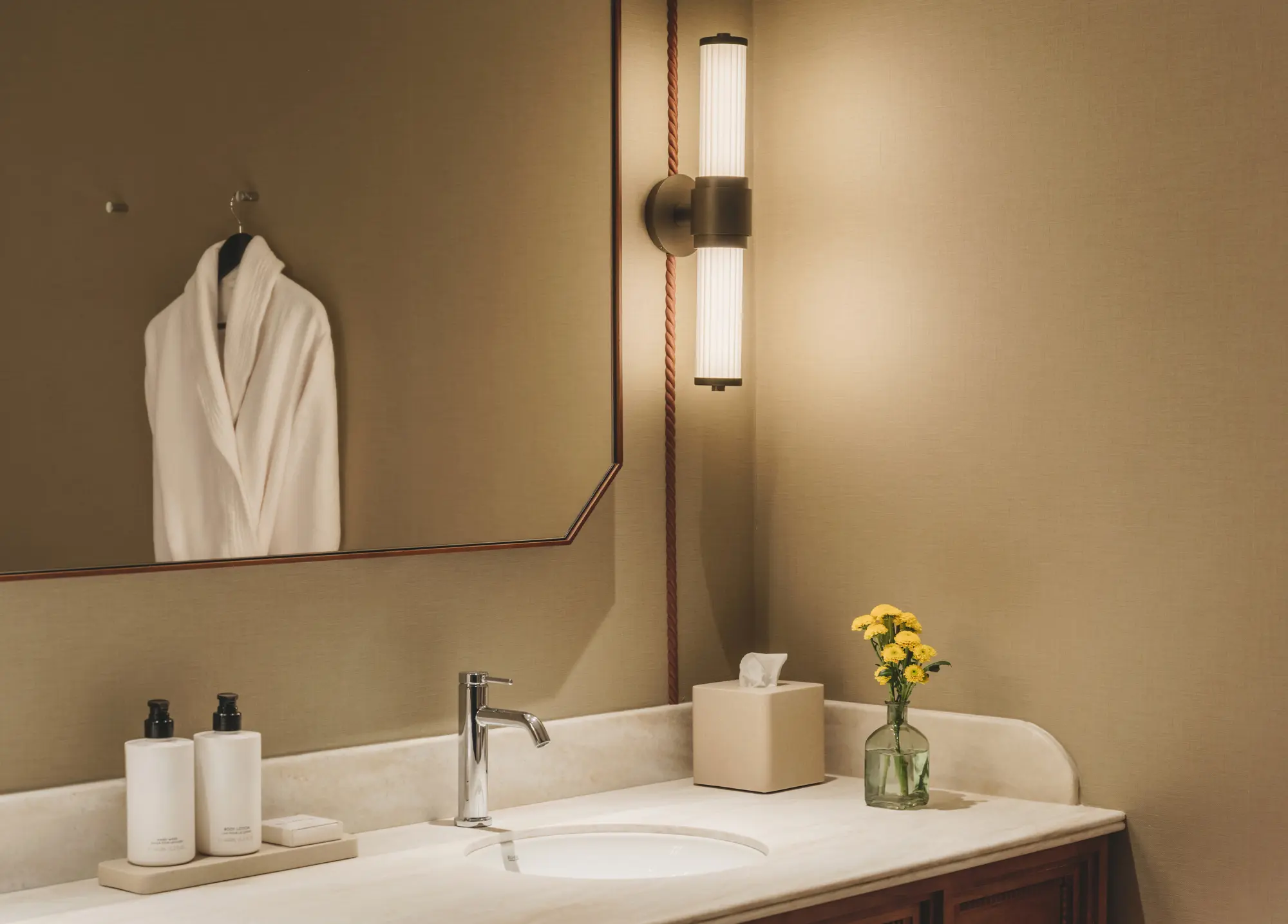 El baño muestra un lavabo con grifo, espejo, toallas dobladas y un jarrón con flores.