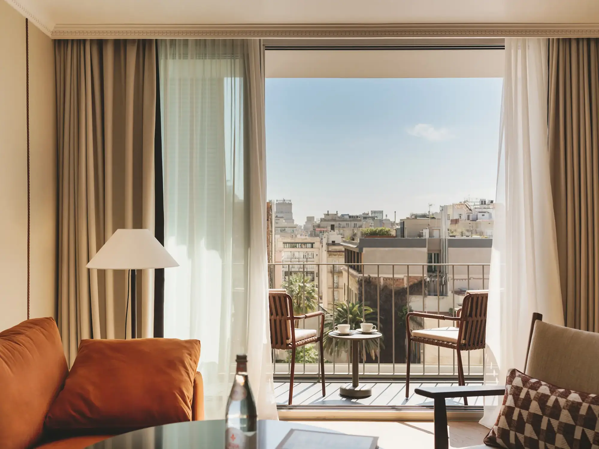 Se ve una sala con sofá, mesa y dos sillas junto a puertas de vidrio que dan a un balcón con vista urbana.
