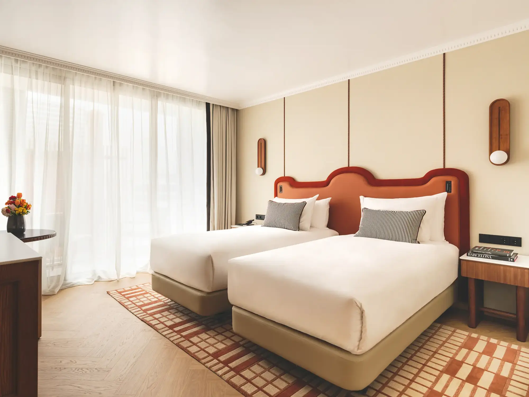 Se ve una cama doble con cabecero de madera, mesitas laterales y cortinas claras junto a una ventana.