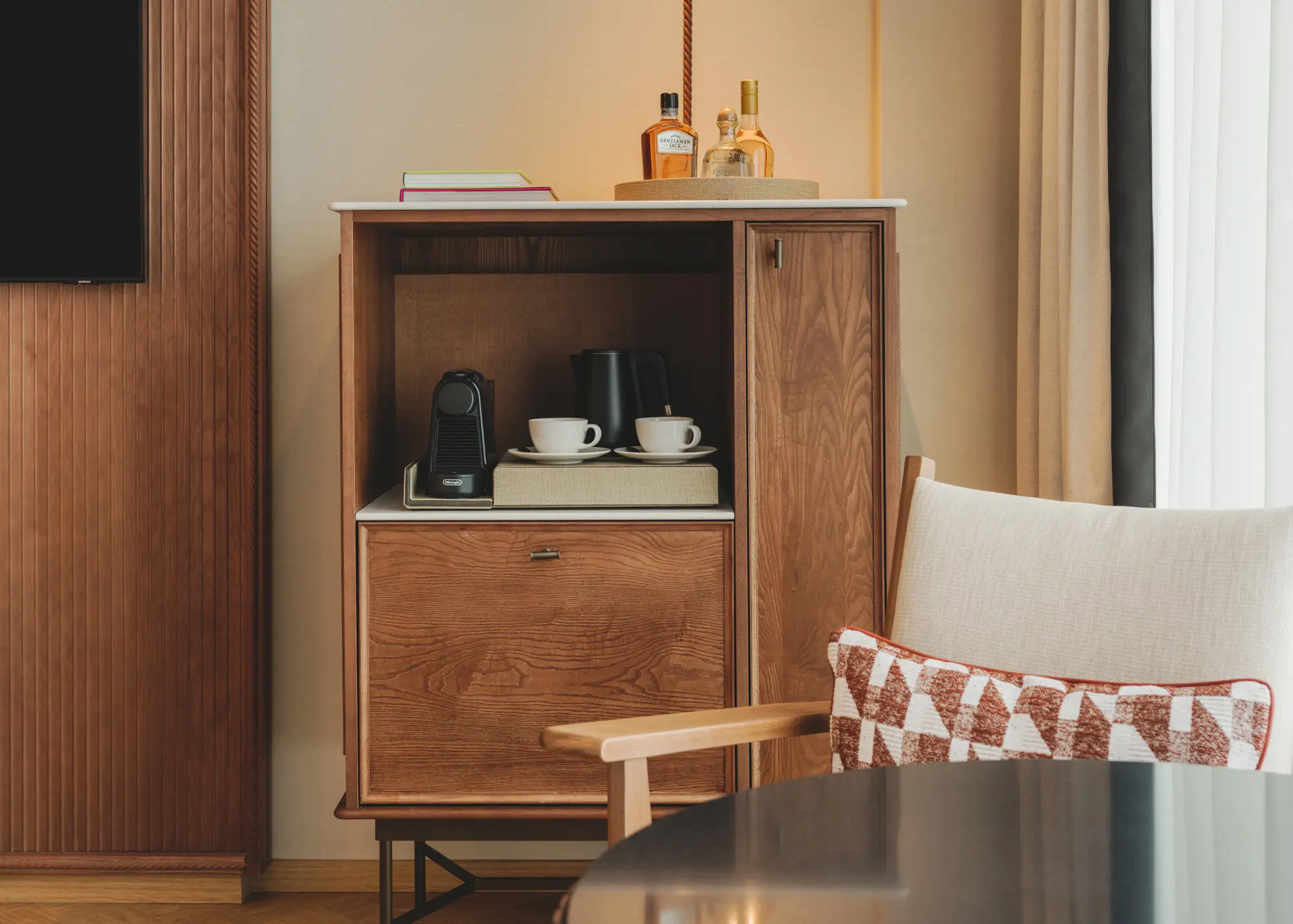 Se ve un mueble de madera abierto con cafetera y tazas, junto a una mesa pequeña y una silla tapizada.