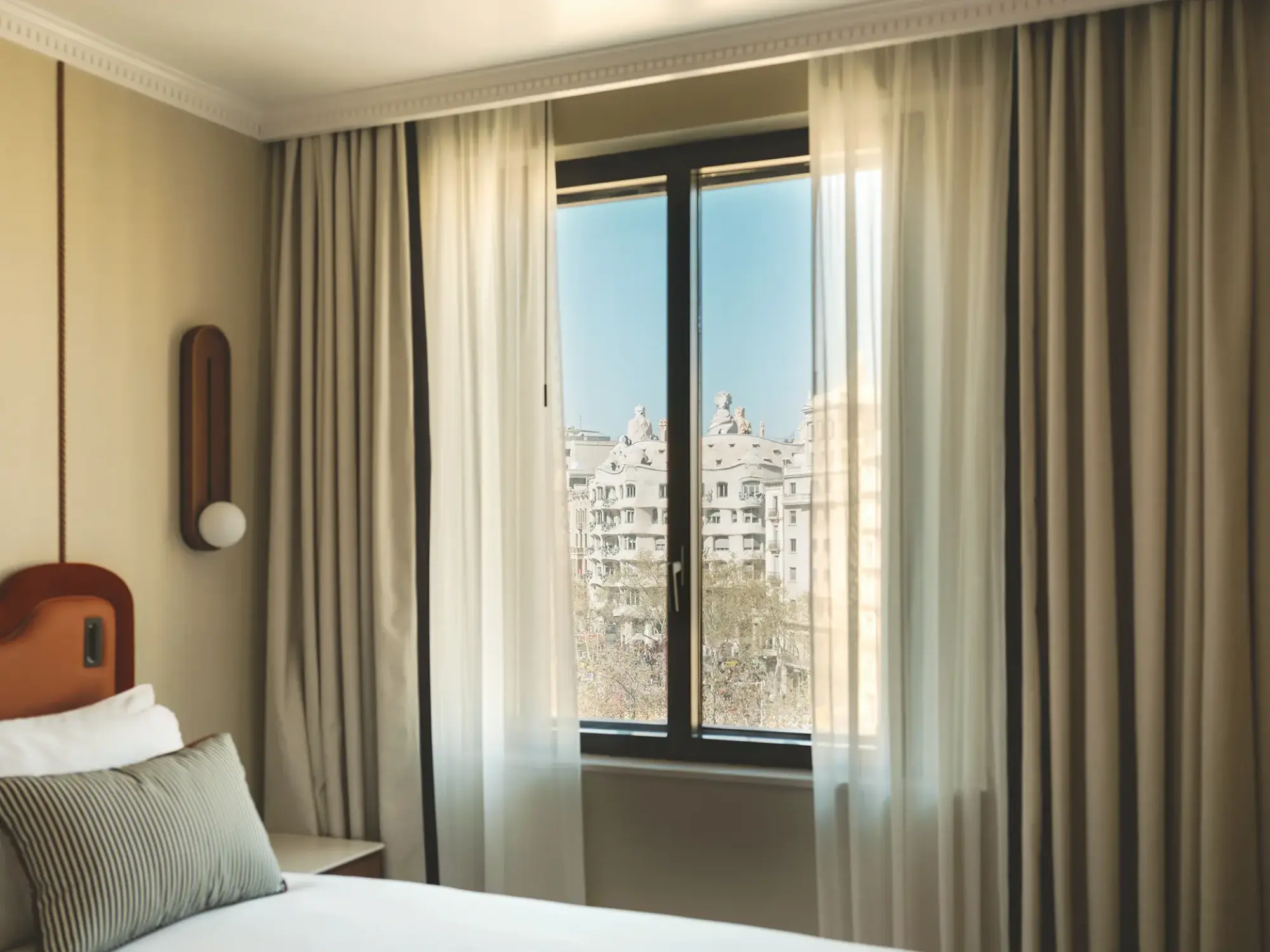 Se ve una cama con almohadas y cortinas junto a una ventana con vista a edificios.