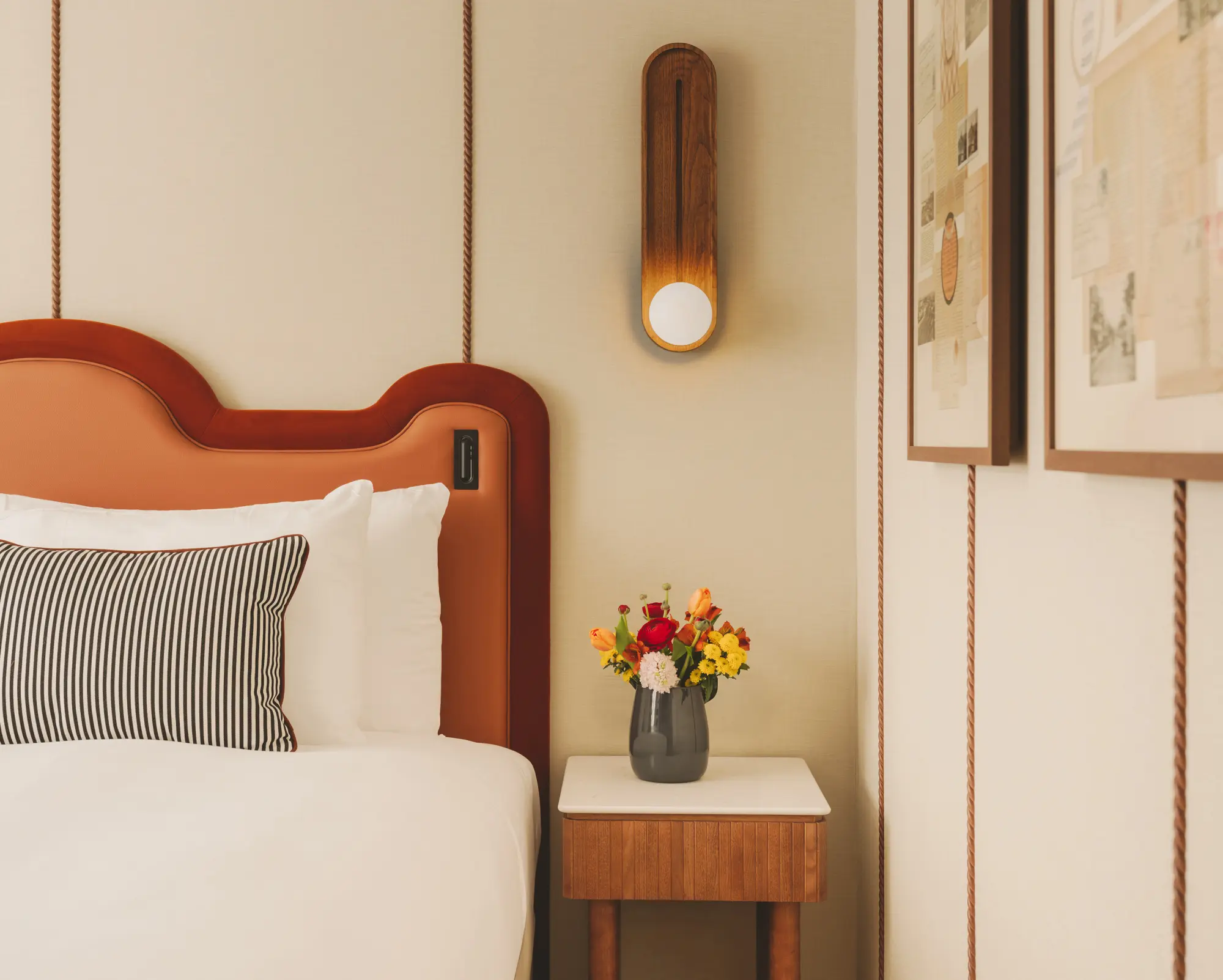 Se ve una cama con cabecero de madera, una mesita auxiliar y un jarrón con flores junto a la pared.
