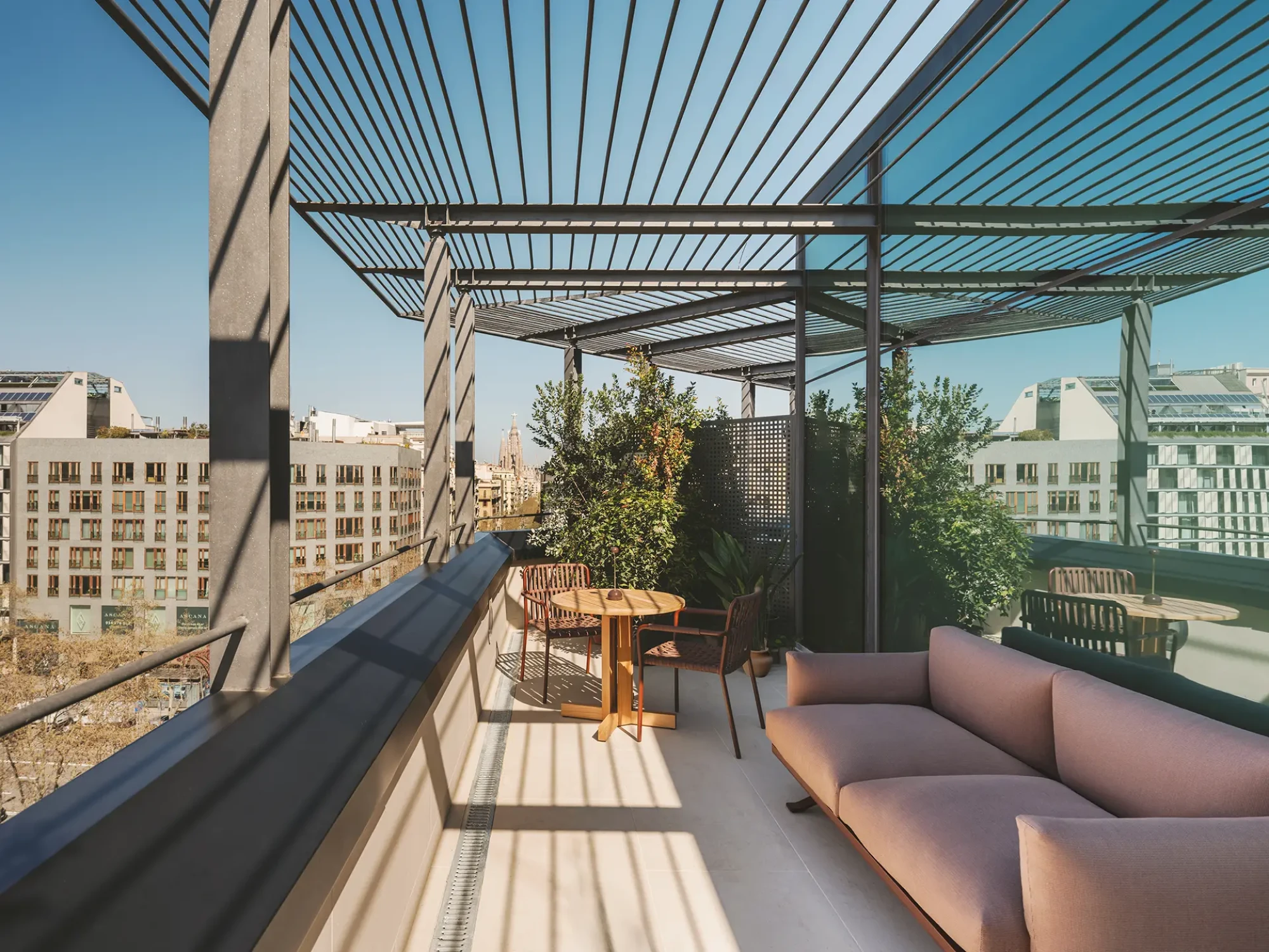 Se ve una terraza con sofá, mesa y sillas bajo una pérgola, con edificios al fondo.