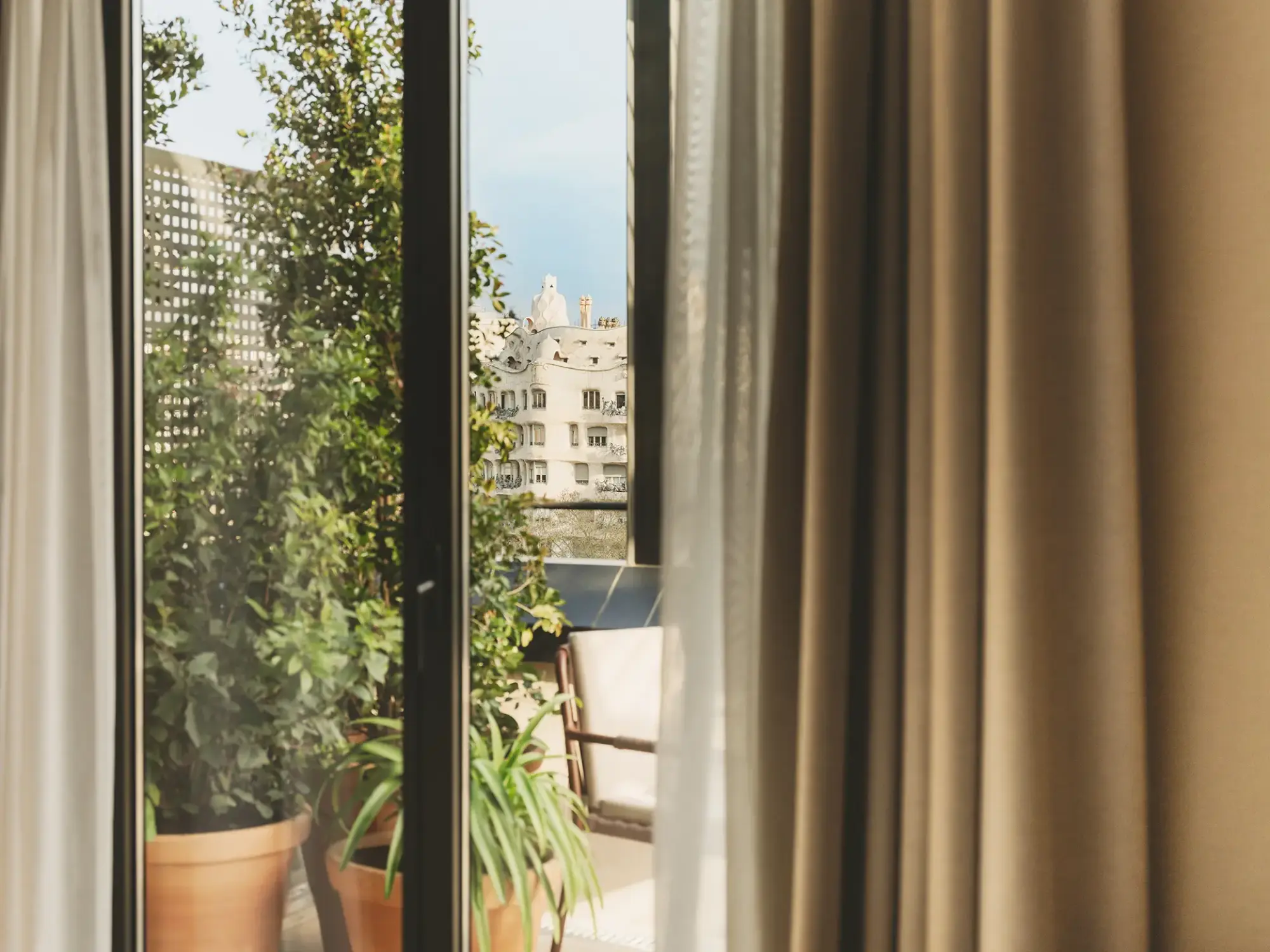 Se ve una terraza con plantas en macetas y una silla junto a una ventana con cortinas.